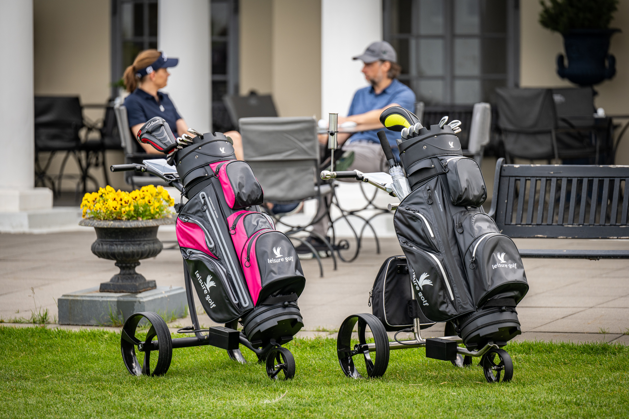 Zwei Golf-Trolleys mit Golfbags stehen auf einer Wiese, im Hintergrund sitzt ein Paar auf einer Terrasse