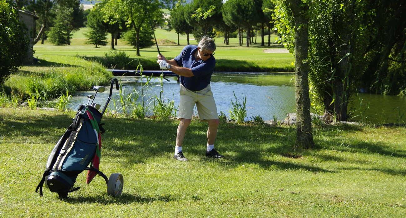 Ein Golfspieler steht auf einer Wiese vor einem kleichen Teich und nimmt Schwung mit einem Golfschläger, neben ihm steht ein Push Golf Trolley.