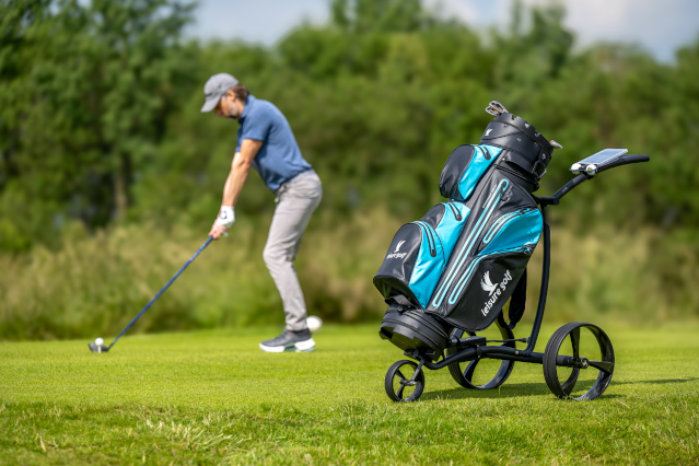 Ein Elektro-Golftrolley mit blau-schwarzer Golfbag steht auf einer Golfwiese, ein Golfspieler ist im Hintergrund mit Schläger.